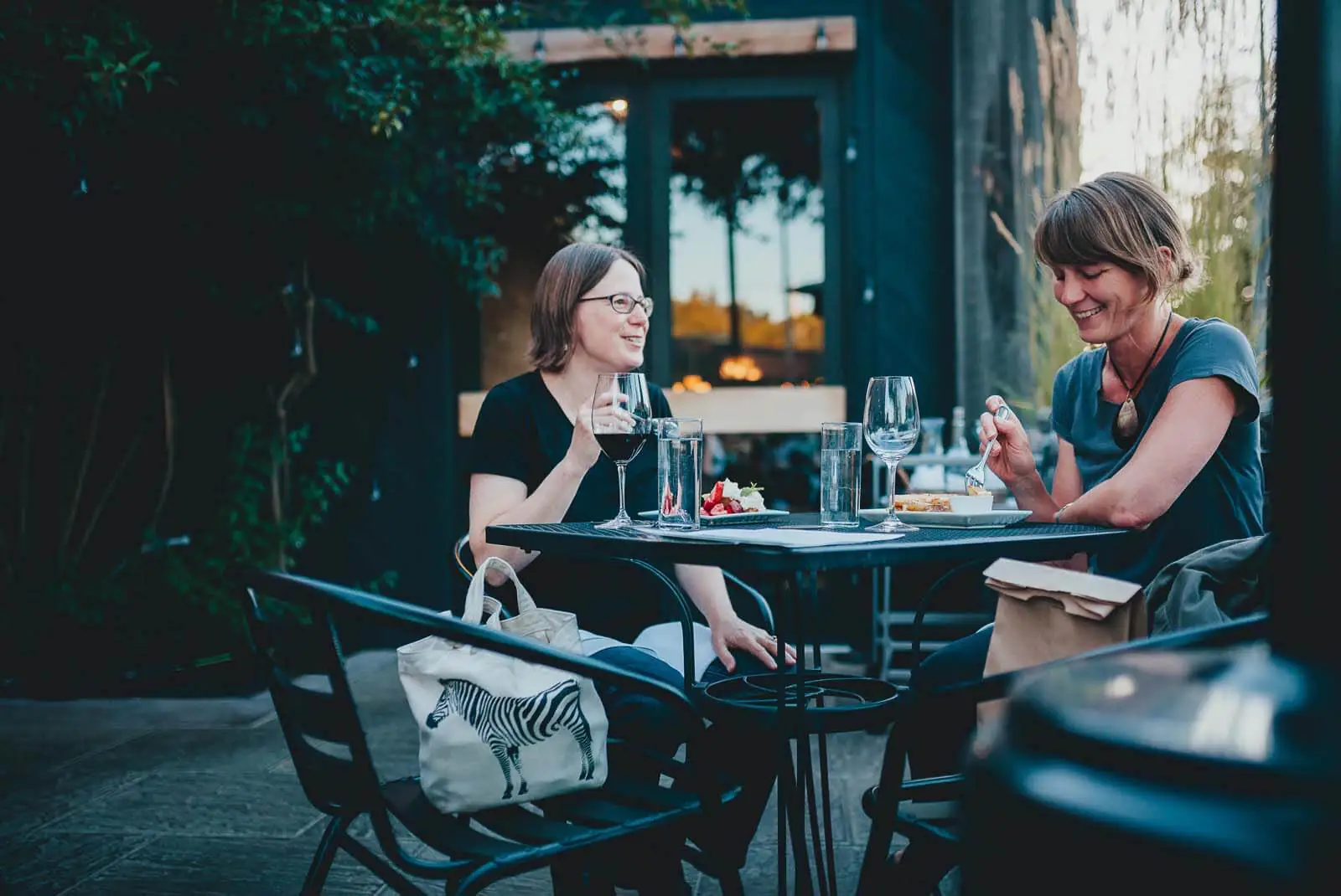 Two women seated at a table outside of a restaurant enjoying food and wine. 